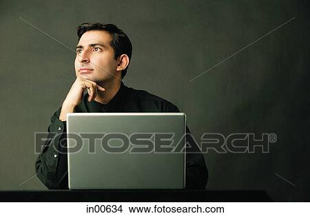 Picture - Man with hand on chin, laptop open in front of him. Fotosearch