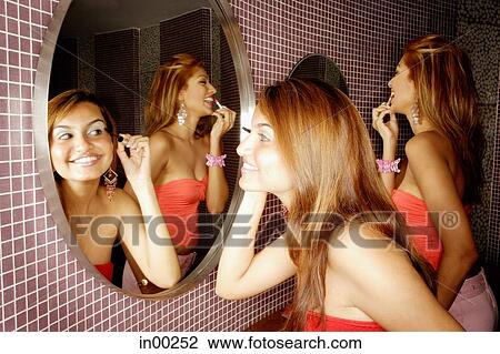 Stock Image - Two women in public restroom, looking at mirror, applying make-up. Fotosearch