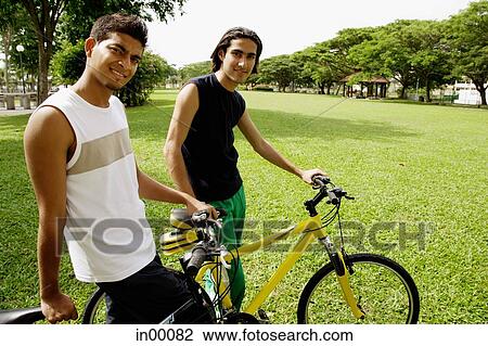 Two young men on bicycles in park, smiling at camera View Large Photo Image Stock Image - Two young men on bicycles in park, smiling at camera. Fotosearch