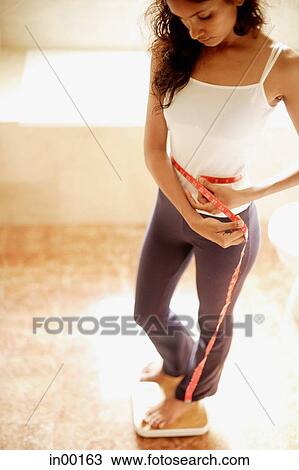 Stock Image - Woman holding tape measure around waist, standing on weight scale. Fotosearch