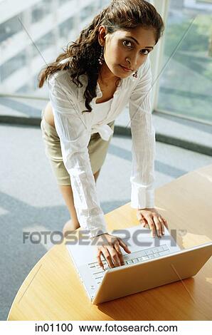 Stock Image - Woman standing over desk, using laptop, looking up at camera. Fotosearch