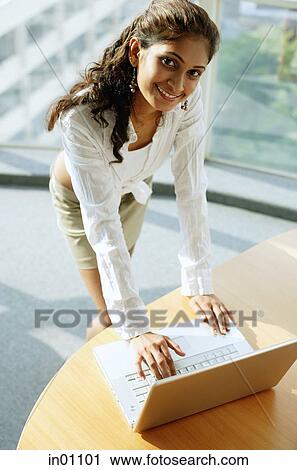 Stock Image - Woman standing over desk, using laptop, looking up at camera. Fotosearch