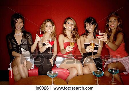 Stock Image - Women sitting side by side, holding drinks up towards camera. Fotosearch