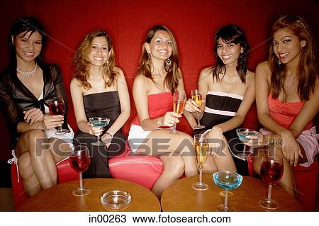 Stock Image - Women sitting side by side, holding drinks, smiling at camera. Fotosearch