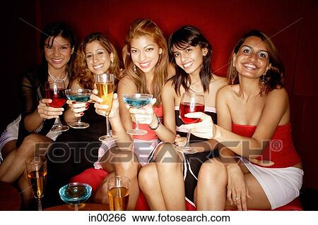 Stock Photograph - Women sitting side by side, smiling at camera, holding drinks. Fotosearch