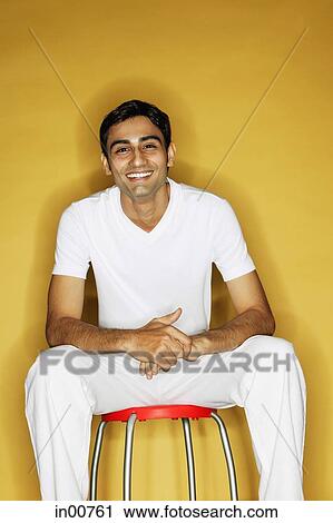 Stock Image - Young man sitting on stool, looking at camera. Fotosearch