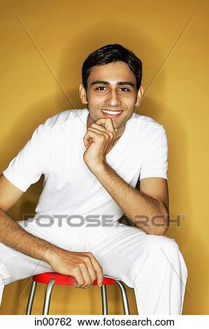 Young man sitting on stool, looking at camera, hand on chin View Large Photo Image Stock Image - Young man sitting on stool, looking at camera, hand on chin. Fotosearch