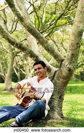 Young man sitting under tree, holding guitar, looking at camera View Large Photo Image Stock Image - Young man sitting under tree, holding guitar, looking at camera. Fotosearch