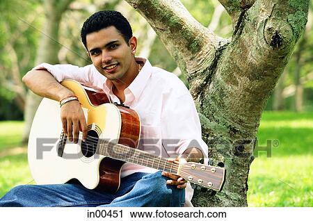 Stock Image - Young man sitting under tree, holding guitar, portrait. Fotosearch