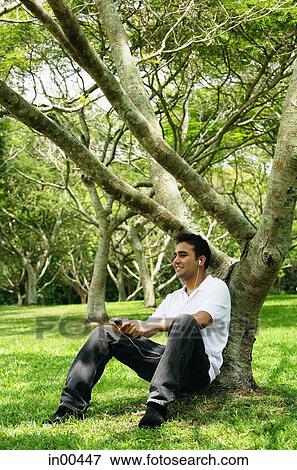 Young man sitting under tree, listening to music View Large Photo Image Stock Photo - Young man sitting under tree, listening to music. Fotosearch