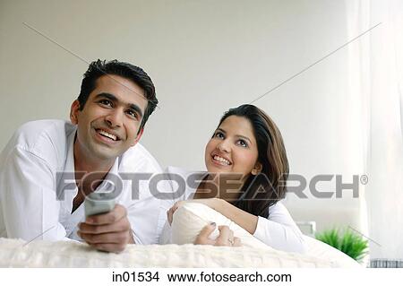 Picture - Couple lying on bed, watching TV. Fotosearch