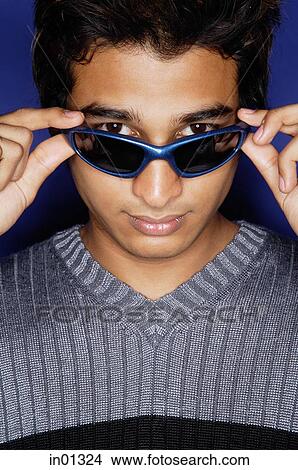 Man against blue background, adjusting sunglasses, looking at camera View Large Photo Image Picture - Man against blue background, adjusting sunglasses, looking at camera. Fotosearch