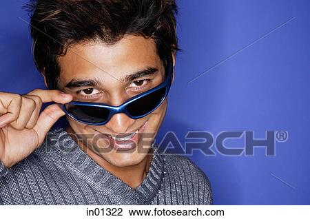 Stock Image - Man against blue background, adjusting sunglasses, smiling at camera. Fotosearch