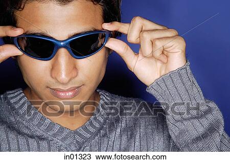 Stock Image - Man against blue background, adjusting sunglasses. Fotosearch