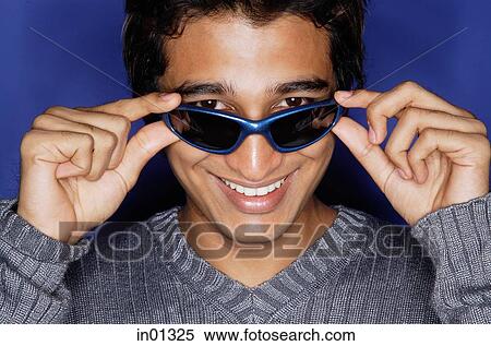 Stock Photography - Man against blue background, adjusting sunglasses, smiling at camera. Fotosearch