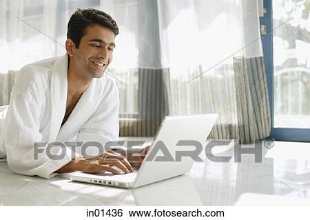 Stock Photograph - Man in bathrobe, lying on floor, using laptop. Fotosearch