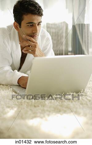 Stock Image - Man in bathrobe, using laptop, hand on chin. Fotosearch