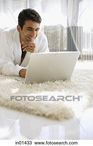 Stock Image - Man in bathrobe, using laptop, hand on chin, smiling. Fotosearch