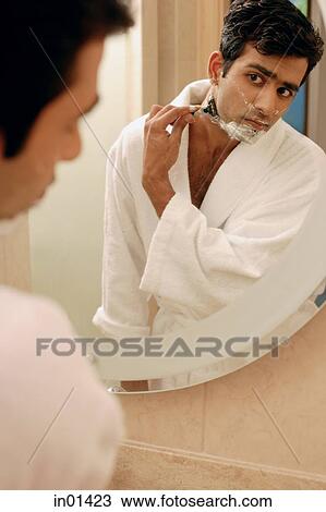 Stock Image - Man looking in bathroom mirror, shaving his face. Fotosearch