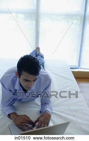 Stock Image - Man lying on bed using laptop. Fotosearch