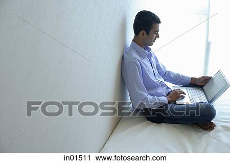 Stock Image - Man sitting on bed, using laptop. Fotosearch