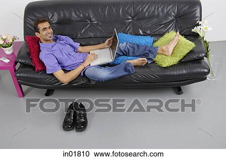 Man sitting on sofa with laptop, smiling at camera View Large Photo Image Stock Image - Man sitting on sofa with laptop, smiling at camera. Fotosearch