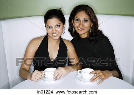 Picture - Two women sitting side by side, having tea, looking at camera. Fotosearch