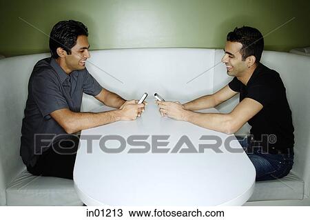 Stock Image - Two young men sitting opposite each other with mobile phones. Fotosearch