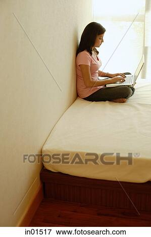 Stock Photo - Woman at home, sitting on bed, using laptop. Fotosearch