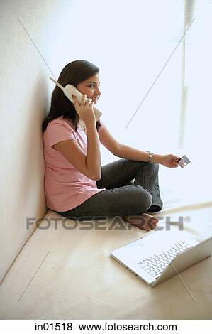Stock Photo - Woman at home, sitting on bed with laptop, using cordless phone. Fotosearch