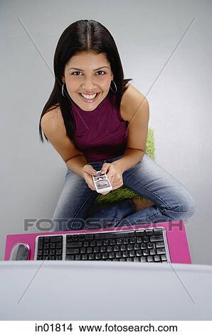 Woman sitting in front of laptop, holding mobile phone, smiling at camera View Large Photo Image Picture - Woman sitting in front of laptop, holding mobile phone, smiling at camera. Fotosearch