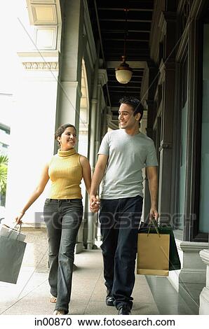 Stock Image - Young man and woman walking, holding hands and carrying shopping bags. Fotosearch