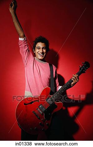 Stock Photo - Young man holding red guitar, standing against red wall, smiling at camera. Fotosearch