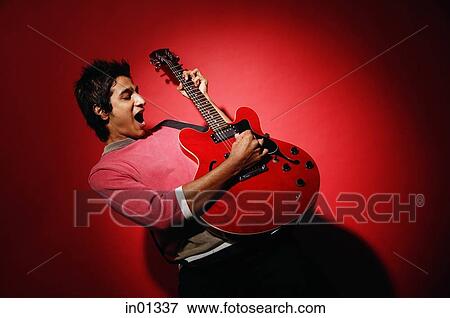Stock Photo - Young man playing guitar, standing against red wall. Fotosearch