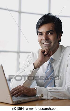 Businessman sitting in front of laptop, hand on chin, looking at camera View Large Photo Image Picture - Businessman sitting in front of laptop, hand on chin, looking at camera. Fotosearch