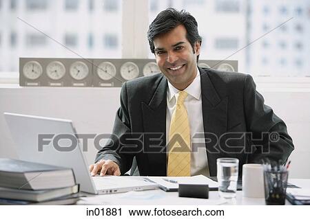 Stock Image - Businessman sitting in office, smiling at camera. Fotosearch