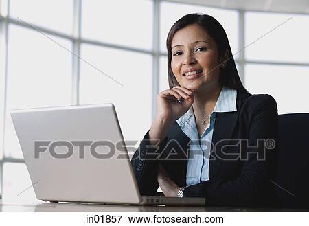 Businesswoman sitting in front of laptop, hand on chin, smiling View Large Photo Image Stock Photo - Businesswoman sitting in front of laptop, hand on chin, smiling. Fotosearch