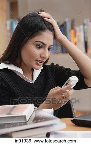 Stock Photograph - Woman in library, hand on head, looking at mobile phone. Fotosearch