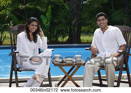 Stock Image - Couple sitting by the pool, looking at camera. Fotosearch