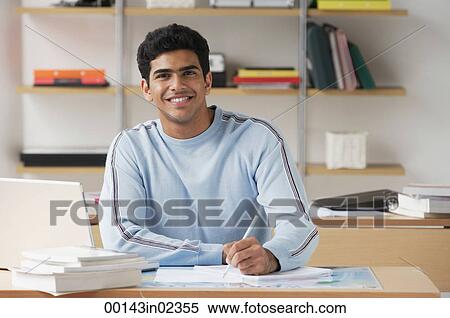 Young man sitting at desk and smiling at camera View Large Photo Image Stock Photography - Young man sitting at desk and smiling at camera. Fotosearch
