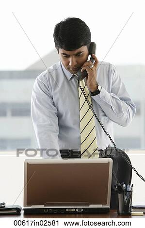 Stock Image - business man standing, using laptop while on phone. Fotosearch