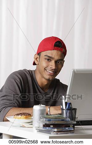 Stock Photo - young man with red cap working on laptop at desk. Fotosearch
