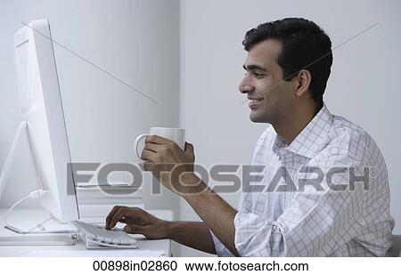 Stock Image - Indian man working on computer and drinking coffee. Fotosearch