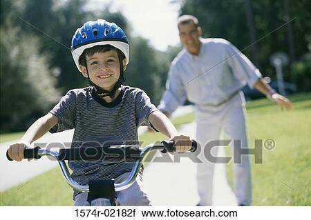 Stock Image - Boy cycling with his father standing behind him. Fotosearch