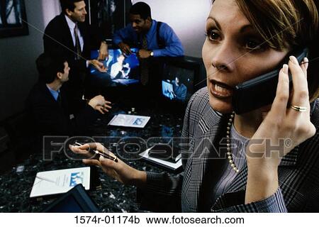Stock Image - Businesswoman talking on a mobile phone with three businessmen talking behind her. Fotosearch