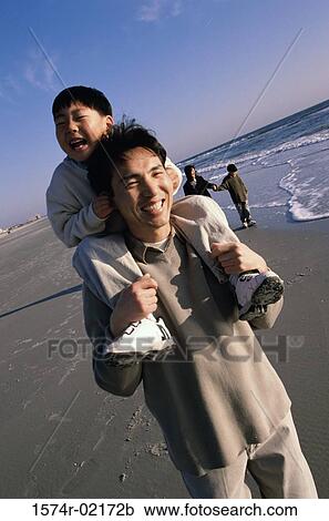 Stock Image - Father carrying his son on his shoulders walking on the beach. Fotosearch