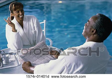 Stock Image - High angle view of a mature couple in bathrobes having breakfast by a swimming pool. Fotosearch