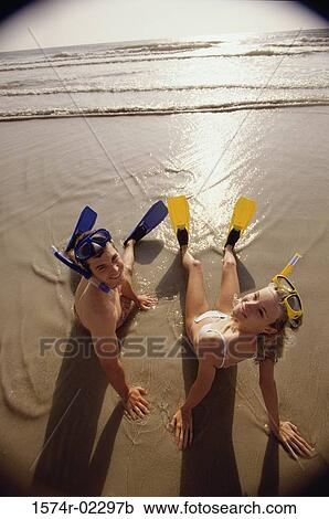 Stock Image - High angle view of a teenage couple sitting on the beach wearing snorkels and flippers. Fotosearch
