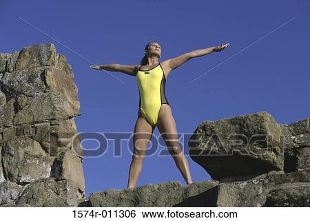 Stock Photograph - Low angle view of a young woman standing on a rock with her arms outstretched. Fotosearch