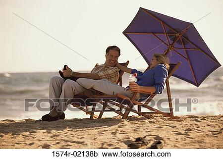 Mid adult couple sitting on lounge chairs on the beach View Large Photo Image Stock Image - Mid adult couple sitting on lounge chairs on the beach. Fotosearch